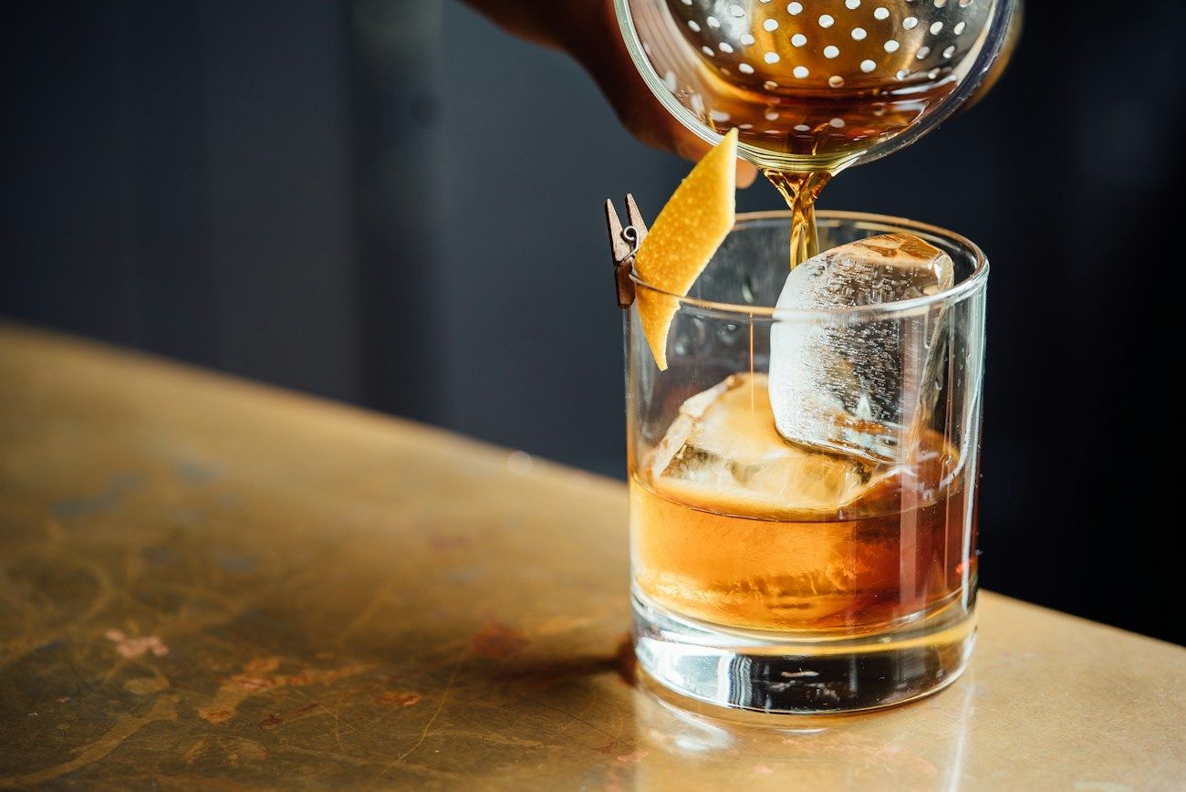 An amber cocktail being poured through a strainer over a large ice cube